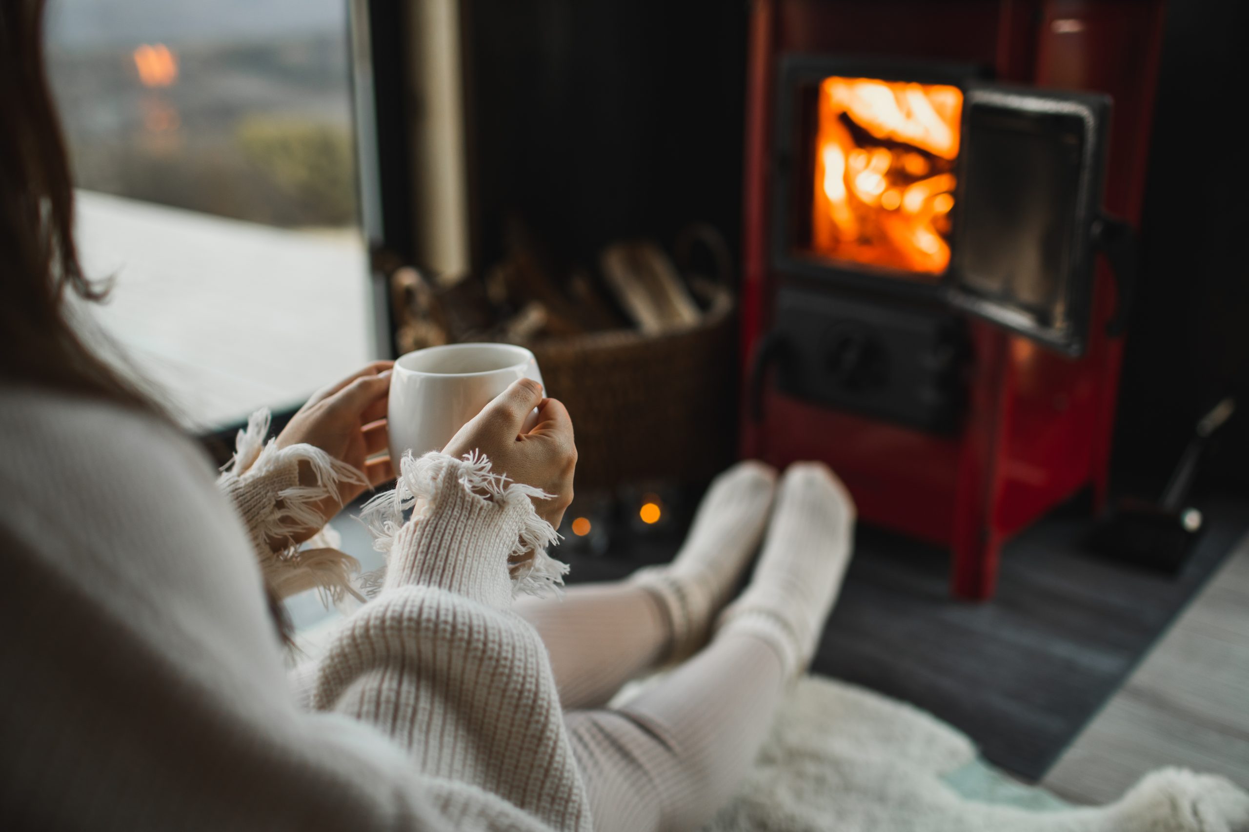 Feet in woollen socks by the Christmas fireplace. Woman relaxing by warm fire with a cup of hot drink and warming up her feet in woollen socks. Cozy atmosphere. Winter and Christmas holidays concept.