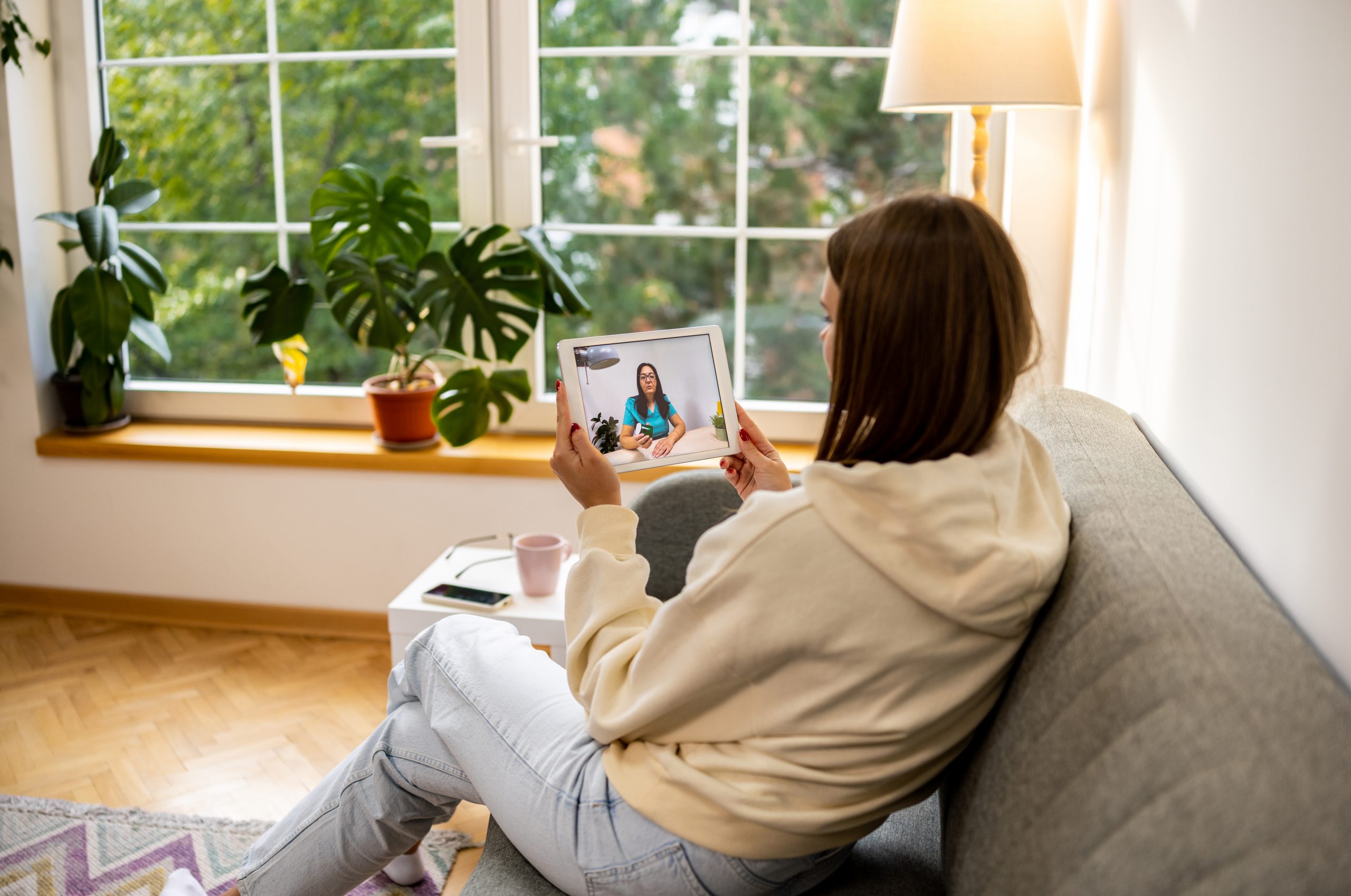 Young woman sitting on couch at her apartment and discussing with her therapist on digital tablet during online session