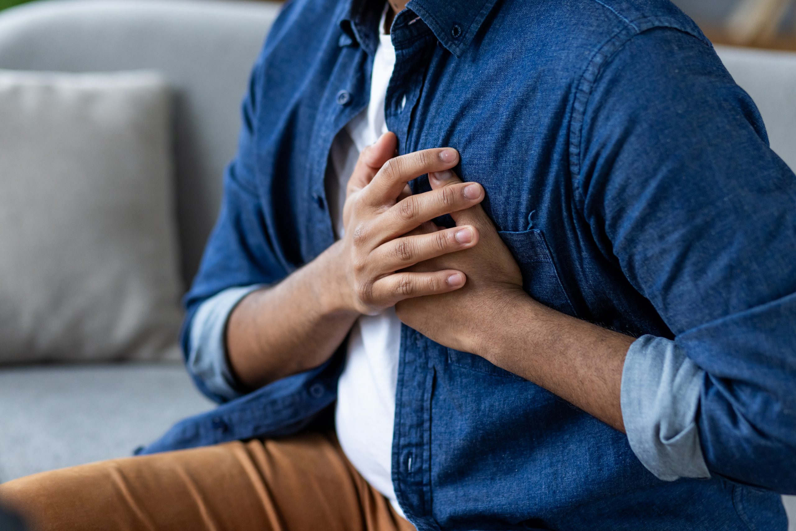 Man holding hands over heart, severe pain, heart attack, sitting at home in living room on sofa close-up.