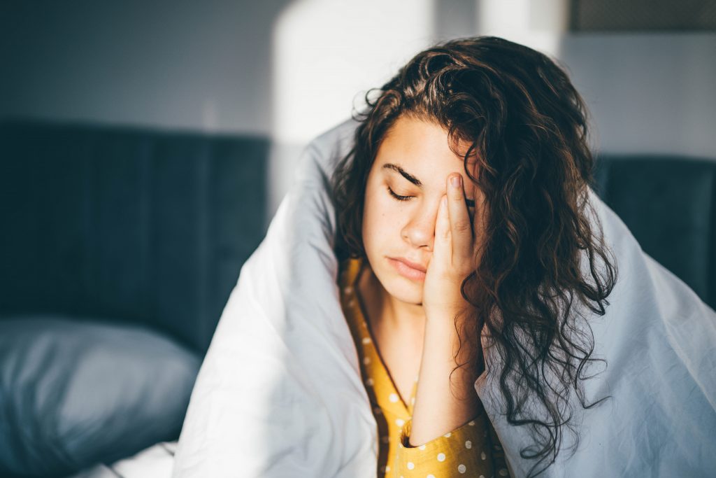 A woman wrapped in a white duvet sits in bed, shielding her eyes from the morning light with a weary expression.